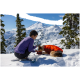 Eine Person in einem lila Langarmshirt kniet im Schnee und füttert einen Hund, der einen orangefarbenen Mantel trägt, vor der Kulisse schneebedeckter Berge.