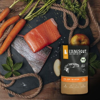 LIEBESGUT-Katzenfutterbeutel mit der Aufschrift "Bio-Lachs für Katzen" neben einem Lachsfilet, Karotten, Apfel und Kräutern auf Schiefer.