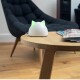 Ein weißes katzenförmiges Gerät mit grünen Ohren sitzt auf einem Holztisch, neben Zeitschriften, in einem modernen Wohnzimmer.