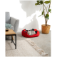 Eine graue Katze, die in einem rot-schwarzen Haustierbett entspannt, in einem gemütlichen Wohnzimmer mit einem grauen Sofa, einem Couchtisch und einer Topfpflanze.