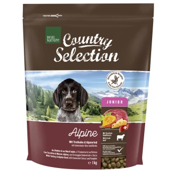 Country Selection Junior Alpine Truthahn & Alpenrind 1 kg Braune Hundefuttertüte mit einem Hund auf der Vorderseite, beschriftet mit "Real Nature Country Selection Alpine," mit Zutaten, die auf Deutsch und Englisch aufgelistet sind.