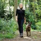Eine Frau in einem schwarzen Outfit führt einen glücklichen, braunen Hund an der Leine durch einen üppigen grünen Waldweg.