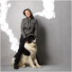 Eine Person in einem grauen Hoodie und schwarzen Hosen sitzt neben einem großen, flauschigen schwarz-weißen Hund, beide vor einem grauen Hintergrund.