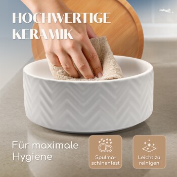 Eine Hand reinigt eine weiße Keramik-Haustierschüssel mit Zickzackmuster, die den Text "HOCHWERTIGE KERAMIK" und "Für maximale Hygiene" zeigt.