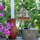 A bird feeder with a small bird perched on it, surrounded by vibrant pink flowers, a clay pot, and a metal watering can.