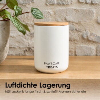Eine getigerte Katze greift in ein weißes Leckerli-Glas mit der Aufschrift 'HANSOME TREATS' auf einem Holztisch, mit einem schwarzen Glas daneben und Kunstwerken an der Wand.