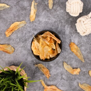 Eine schwarze Schüssel, gefüllt mit Trockenfischhaut-Leckerlis, umgeben von verstreuten Stücken, einem geflochtenen Korb mit grünem Gras und zwei weißen Seilspielzeugen.