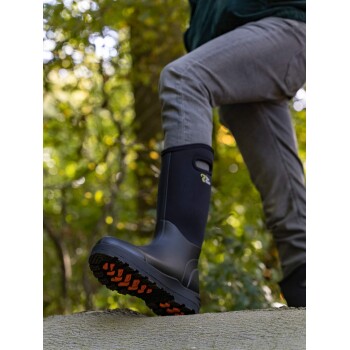 Eine Nahaufnahme eines Beins einer Person, die einen schwarzen Gummistiefel mit einer orangefarbenen Sohle trägt, während sie auf einen Baumstamm in einem grünen, verschwommenen Waldhintergrund tritt.