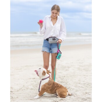 Eine Frau in einem weißen Shirt und Jeansshorts steht am Strand, hält einen rosa Ball und eine Hundeleine. Ein brauner und weißer Bulldog schaut aufgeregt zu ihr auf.
