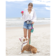 Eine Frau in einem weißen Shirt und Jeansshorts steht am Strand, hält einen rosa Ball und eine Hundeleine. Ein brauner und weißer Bulldog schaut aufgeregt zu ihr auf.