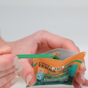 Eine Hand, die eine grüne Schaufel hält und ein braunes Pulver aus einer JBL Pro Terra Verpackung entnimmt, beschriftet mit "CRUNCHY."