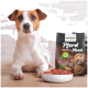 Ein weißer Hund liegt auf Holz neben einer Schüssel mit roten Fleischstücken und einem proCani Pferd-Menü-Hundefutterpaket mit einem braunen Hund darauf.