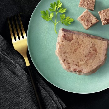 Une tranche de pâté pour animaux de compagnie sur une assiette vert clair, accompagnée de petits cubes du même pâté et d'une branche de persil, avec une fourchette dorée à proximité.
