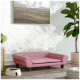 Ein stilvolles pinkes Haustiersofa mit Knopfdetails steht auf einem Holzboden, neben einer weißen Futterschüssel, die mit Trockenfutter gefüllt ist, und einer Topfpflanze.