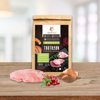 Eine Tüte Wildfang-Hundefutter mit der Aufschrift "Truthahn mit Süßkartoffel & Preiselbeere" liegt auf Holz mit rohem Truthahn, Süßkartoffeln und Preiselbeeren.