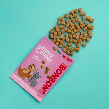 Un sac de friandises pour animaux de compagnie rose étiqueté "edgard Cooper" avec des images de dessins animés d'un chien et d'un lapin, entouré de petites friandises brunes sur un fond bleu-vert.