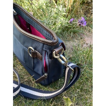 Eine blaue Tragetasche für Haustiere mit pinkem Innenfutter, mit braunen Lederakzenten und einem gestreiften Tragegurt, die auf Gras neben Lavendelblüten ruht.