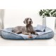 Ein grauer Weimaraner-Hund, der auf einem plüschigen blauen Haustierbett mit einem kleinen passenden Kissen entspannt, vor einem hellen Hintergrund mit einer Pflanze.