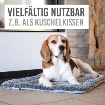 Ein Beagle, der auf einer weichen, grauen Tiermatte in einem modernen Innenraum liegt. Der Text darüber lautet "Vielfältig nutzbar z.B. als Kuschelkissen."