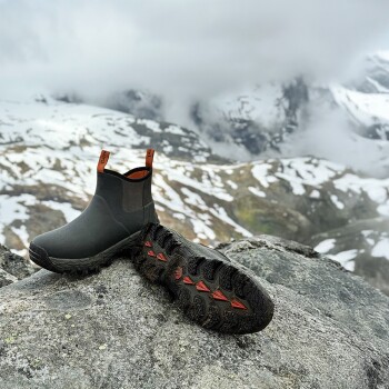 Schwarze und orangefarbene Hundestiefel mit robusten Sohlen auf einem Felsen, verschneiter Bergkulisse, beschriftet mit „Gaiter“ und „Tire“.