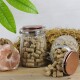 Zwei Glasbehälter mit braunen Tierpellets, einer aufrecht und einer umgekippt, sodass Pellets auf Stroh fallen, mit kupferfarbenen Deckeln und grünen Blättern darüber.