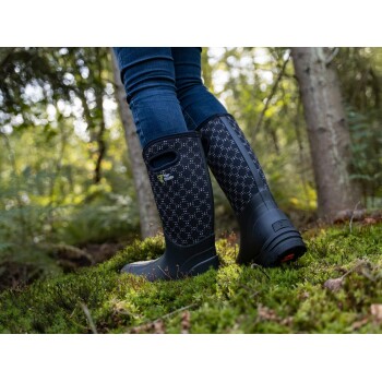 Eine Person in stilvollen schwarzen gemusterten Gummistiefeln steht auf üppigem grünen Moos in einem Wald, mit Bäumen und sanftem Sonnenlicht im Hintergrund.
