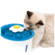 Un chat crémeux et duveteux avec des marques sombres boit dans une fontaine à eau pour animaux bleue et blanche en forme de fleur, avec de l'eau qui coule.