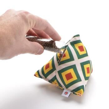 Eine Hand, die braune Haustierleckerlis aus einem Glasrohr in ein buntes, gemustertes, dreieckiges Stoffspielzeug für Haustiere gießt.