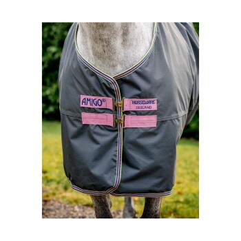 Ein graues Pferd, das eine Amigo Horseware Ireland Decke mit pinken Schnallen und buntem Besatz trägt, steht auf Gras.