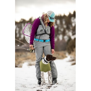 Eine Frau in einem lila und grauen Angeloutfit steht im Schnee, hält Angelruten, mit einem braun-weißen Hund in einem grünen Pullover neben ihr.