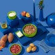 Ein blauer Tisch zeigt eine grüne Schüssel mit Tierfutter, umgeben von Süßkartoffeln, Hüttenkäse, einem Pfirsich, Öl und einer blauen Kerze.