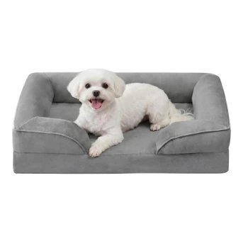 Kleiner weißer Malteser-ähnlicher Hund liegt in einem grauen, gepolsterten rechteckigen Haustierbett, blickt in die Kamera, mit herausgestreckter Zunge und fröhlichem Ausdruck.