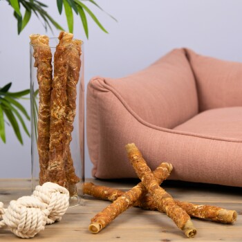 Eine Glasvase, gefüllt mit verdrehten Hundekauartikeln, steht neben einem rosa Hundebett und einem Seilspielzeug auf einer Holzoberfläche.