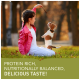 Ein Mädchen in einer rosa Jacke sitzt auf Gras und hält ein Leckerli über einem aufgeregten Hund. Text: "PROTEINREICH, NÄHRSTOFFBALANCIERT, LECKER!"