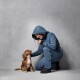 Eine Person in einem blauen Hoodie hockt sich nieder und interagiert mit einem freundlichen Cavalier King Charles Spaniel vor einem neutralen grauen Hintergrund.