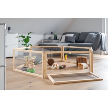 Une enceinte en bois pour animaux de compagnie avec deux cobayes, des jouets et des gamelles, située dans un salon moderne avec un canapé gris et des plantes.