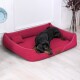 Ein schwarzer Dobermann liegt bequem auf einem lebhaft roten Hundebett mit einem kleinen Kissen, vor einem hellen Holzboden und einer weißen Wand.