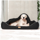 Ein großer weißer Hund, der bequem in einem schwarzen Haustierbett mit einer gemütlichen Decke, die teilweise darauf liegt, vor einer hellen Wand ruht.