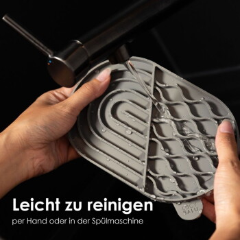 Eine Person hält eine graue Futtermatte für Haustiere unter fließendem Wasser, mit dem Text "Leicht zu reinigen per Hand oder in der Spülmaschine."