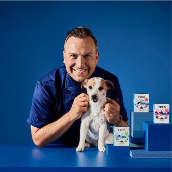 Eine Person in einer blauen Kochjacke hält einen kleinen Hund, während drei bunte Haustierfutterboxen mit der Aufschrift "FRED" auf blauen Blöcken präsentiert werden.