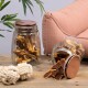 Ein Glasgefäß gefüllt mit Haustierleckereien, einige fallen auf einen Holztisch, neben zwei weißen Seilspielzeugen und einem rosa Sofa im Hintergrund.