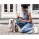 Eine Frau in einem Jeans-Overall kniet neben einem weißen Bulldoggen auf einer Kopfsteinpflasterstraße und hält einen grünen Hundekotbeutelspender.