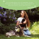Eine Frau sitzt in einem Garten mit lila Blumen und hält eine schwarze Katze neben einem großen Hund, begeistert von den Wolfsbacher Natur-Haustierprodukten.