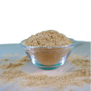 Eine klare Glasschüssel gefüllt mit hellbraunem Pulver, umgeben von verstreutem Pulver auf einer weißen Oberfläche, vor einem grünen Hintergrund.