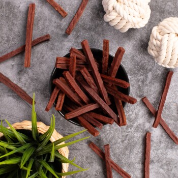 Eine schwarze Schüssel, gefüllt mit langen, dünnen Hundeleckerlis, steht auf einer grauen Oberfläche, umgeben von verstreuten Leckerlis und zwei weißen Seilspielzeugen.