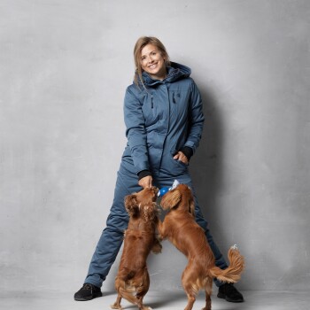 Eine Frau in einem blauen Overall spielt mit zwei kleinen braunen Hunden und beschäftigt sie mit einem blauen Spielzeug, vor einem grauen Hintergrund.