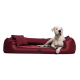 Ein weißer Hund, der auf einem plüschigen burgunderfarbenen Hundebett mit einem passenden Kissen liegt, vor einem schwarzen Hintergrund.