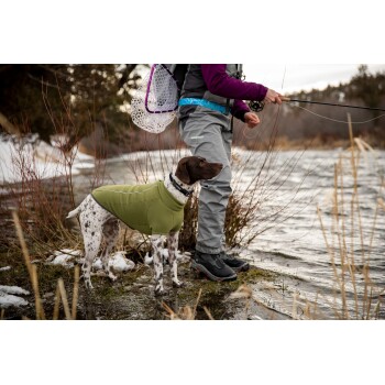 Eine Person in grauen Angelstiefeln steht am Fluss, hält eine Angelrute und hat einen braun-weißen Hund in einem grünen Mantel neben sich.
