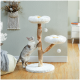 Eine verspielte grau-weiße Katze klettert auf einen flauschigen, blumenförmigen Katzenbaum mit zwei Plattformen und baumelnden Pompons, der in einem gemütlichen Raum steht.