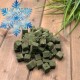 Haufen grüner, gefriergetrockneter Futterwürfel auf einem Holzbrett, rechts eine Pflanze, blaues Schneeflocken-Label mit der Aufschrift 'Gefrier-getrocknet'.
